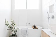 a minimalist airy bathroo with white tiles everywhere, a wooden vanity, a bathtub, a potted plant and a white sink