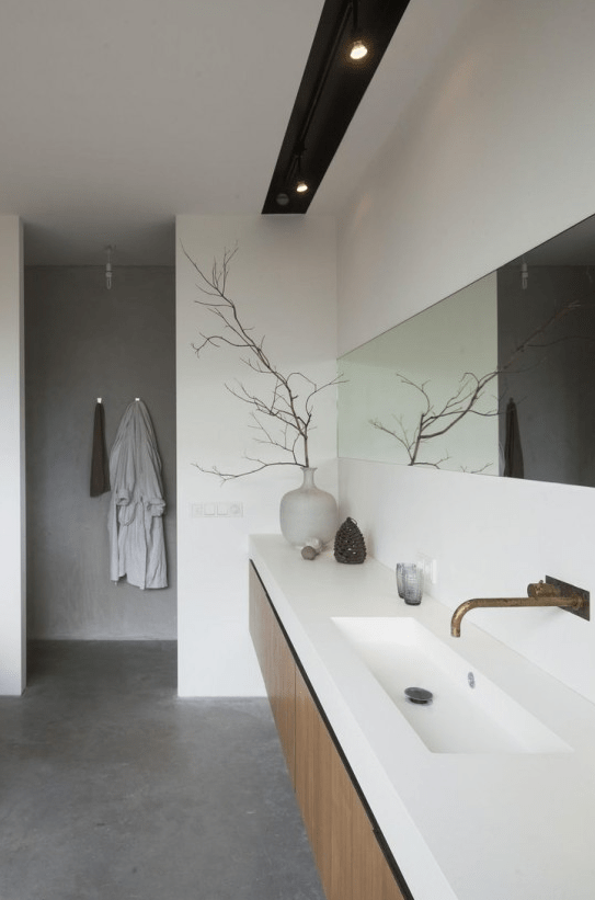 a minimalist bathroom done with white concrete, with a grey concrete floor, a long wooden vanity and a long and narrow mirror plus lights over it