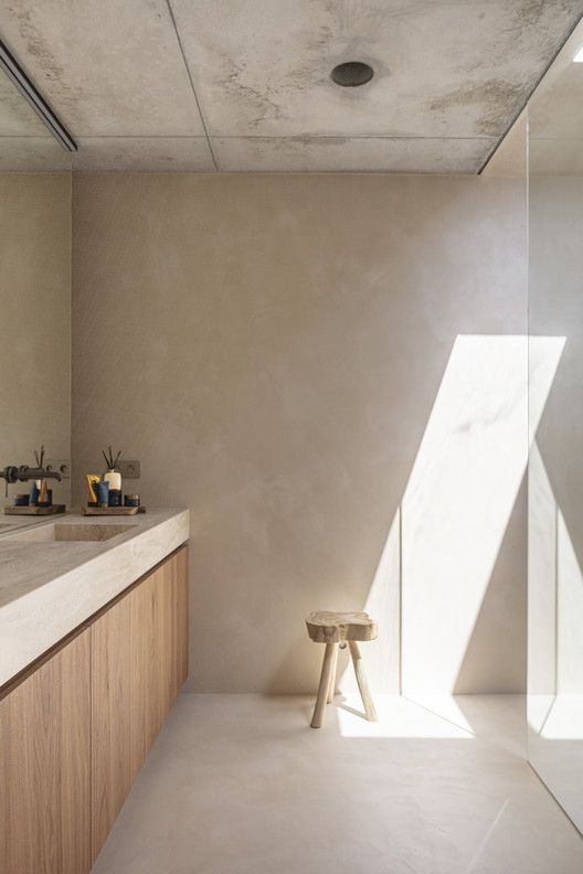 a minimalist bathroom with tan walls, a shower space, a stained vanity with sinks, brass fixtures and a small stool