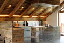 a minimalist chalet kitchen done with sleek wooden cabinets, a small kitchen island, a large hood with lights and built-in lights