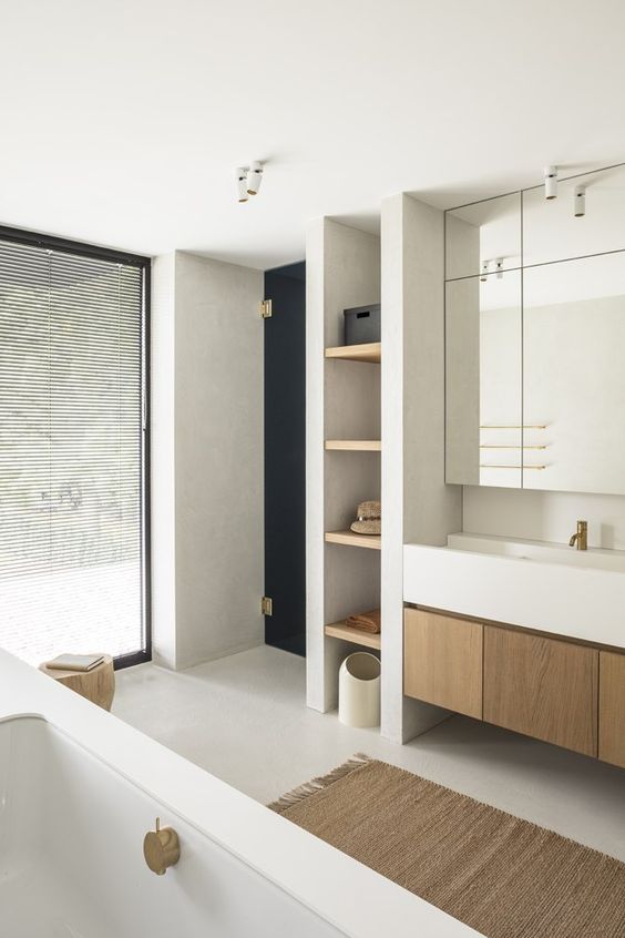 A minimalist concrete bathroom with a tub, a built in stained vnaity, a sink, a mirror cabinet, shelves and a window covered with shades