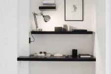 a minimalist home office nook in white, with black shelves and a floating desk, a lamp, artworks and a black chair