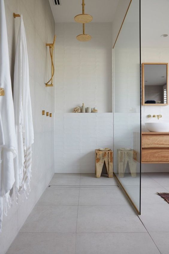 a minimalist white bathroom done with stacked tiles, a stained vanity, a stool and gold fixtures is cool and cozy
