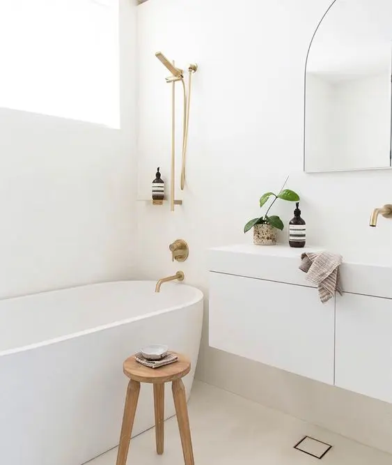 a minimalist white bathroom with a tub, a white floating vanity, an arched mirror, a stool and gold fixtures