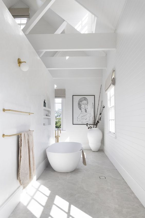 a minimalist white bathroom with a tub, windows that fill the space with light, artwork and gold fixtures