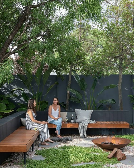 a modern backyard with a long corner wooden bench, a fire pit and lots of plants all around