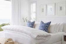 a modern beach bedroom in white, with a large bed, a storage bench and printed blue pillows plus a gorgeous sea view
