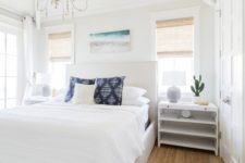 a modern beach bedroom with a jute rug, a white bed and nightstands, a vintage chandelier and a bright artwork
