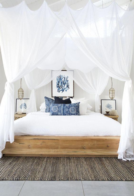 a modern beach bedroom with a wooden bed, a jute rug, printed blue pillows, sea artworks and airy curtains plus Moroccan lamps