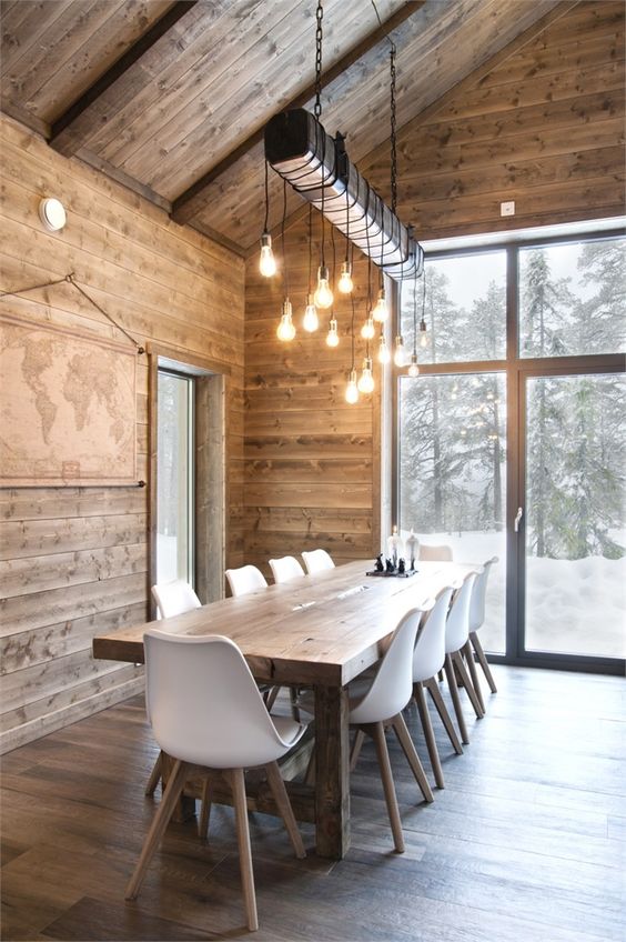 a modern chalet dining space with a wooden table, white chairs, an artwork and a creative lamp of bulbs