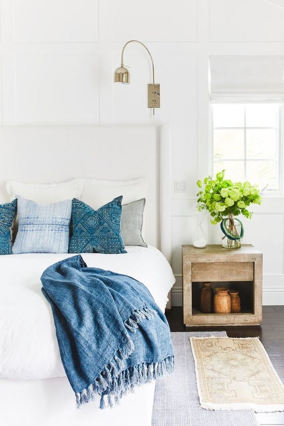 a modern coastal bedroom in neutrals, with a wooden nightstand, layered rugs, greenery and printed blue bedding for a coastal feel