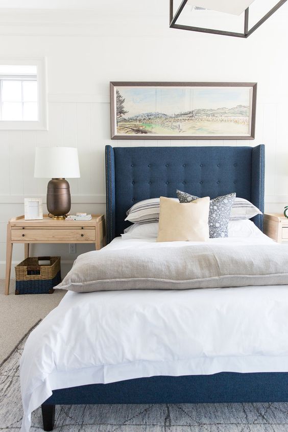 a modern coastal bedroom with a blue bed, printed textiles, a large artwork, wooden furniture, a basket for storage