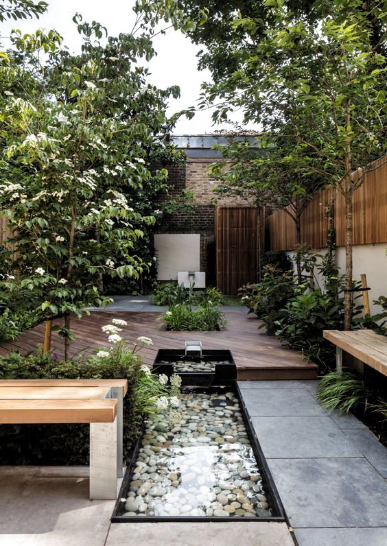 a modern natural backyard with a deck, wooden benches, a couple of waterfalls with pebbles and greenery