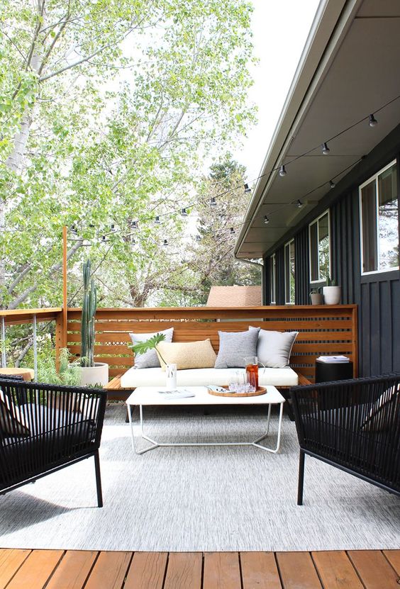 a modern patio with simple wooden and woven furniture, a white table, a grey rug and potted greenery and plants