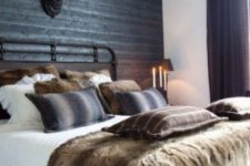 a moody chalet bedroom with a black wood clad wall, a vintage forged bed, faux fur pillows and black lamps