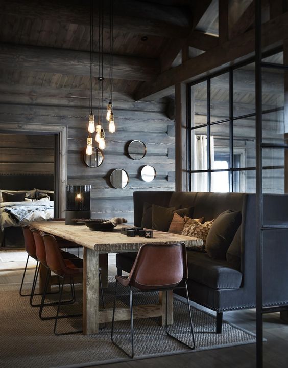 a moody chalet dining space with a large black sofa, pendant bulbs, a wooden table and leather chairs