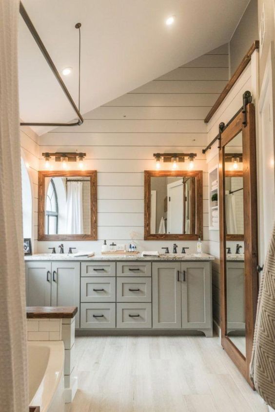 a neutral farmhouse bathroom with white beadboard, grey furniture, wood frame mirrors, sconces and a tub