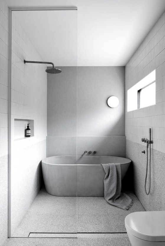 A simple minimalist bathroom clad with skinny white tiles and grey stone like ones, a concrete tub, white appliances