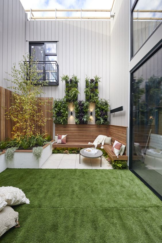 a simple modern backyard with a green lawn, a built in wooden bench, greenery under the bench and on the walls and pillows