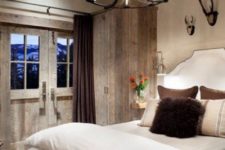 a vintage chalet bedroom done with reclaimed wood, an upholstered bed, a vintage chandelier and faux antlers on the wall