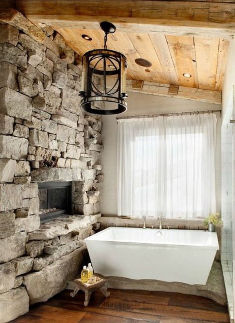 a welcoming chalet bathroom with a wooden floor and ceiling, rock clad walls, a fireplace, a geo tub and a metal lamp