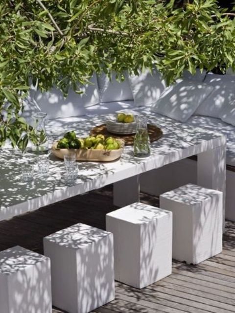 a white minimalist terrace with a dining space, a concrete bench, table and stools and lots of greenery