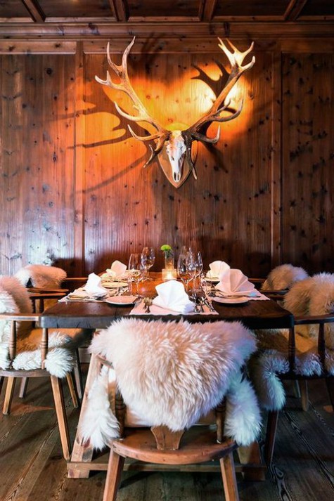 an eclectic chalet dining space with a vintage table and rustic chairs, a deer head and faux fur on the chairs