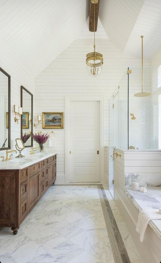 an elegant vintage farmhouse bathroom with white beadboard, a large wooden vanity and mirrors plus cool gold touches