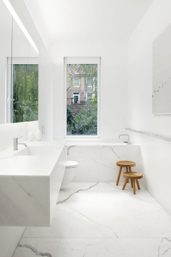 an exquisite white minimalist bathroom fully done with white marble including marble cladding the bathtub, a window and a statement artwork