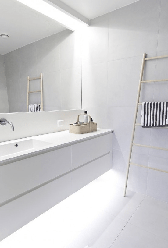 an ultra-minimalist white bathroom with a floating vanity with built-in lights, a statement mirror and a ladder for storing towels