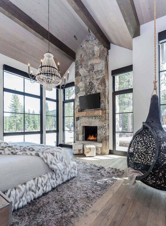 an ultra modern chalet bedroom with a stone fireplace, a suspended chair, a catchy chandelier and lots of faux fur