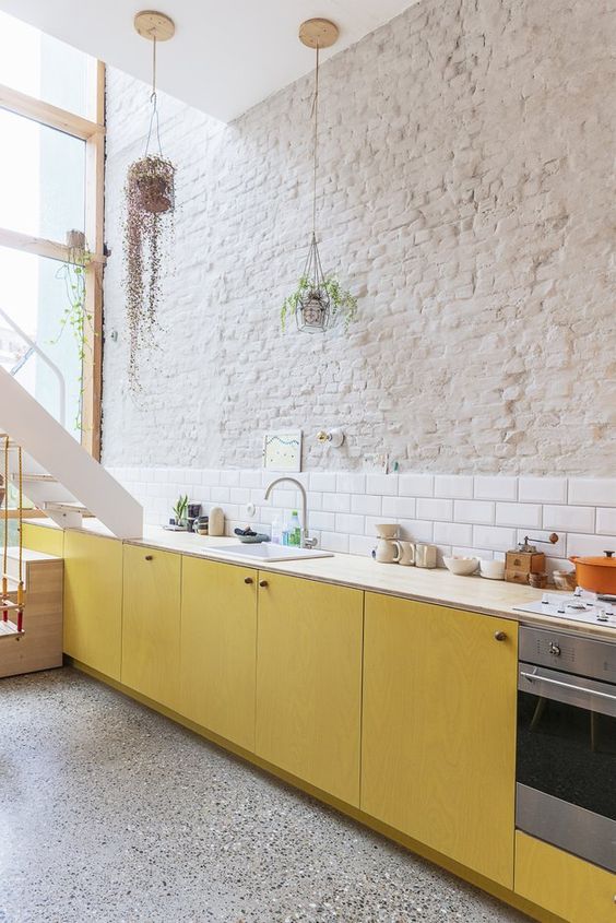 a contemporary kitchen with a brick wall and a white tile backsplash plus yellow plywood cabinets
