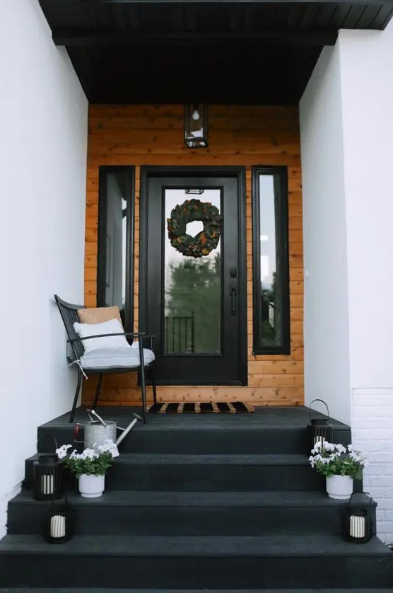 a bold modern porch with a black floor and steps, a black chairs with pillows, potted blooms and candle lanterns and a wreath
