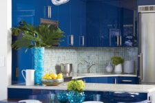 a bright blue kitchen with sleek and shiny cabinets, a neutral tile backsplash and pendant lamps plus white countertops