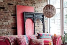 a bright boho living room with a brick wall, a hot pink sofa and ottoman, a wicker lamp and a glazed wall