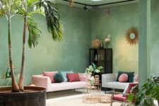 a bright living room with green plaster walls and a ceiling, with grey and pink furniture and colorful pillows and lots of potted greenery