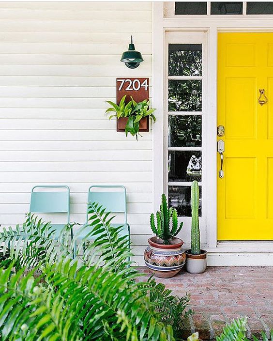 a bright modern porch with a sunny yellow door, cacti in pots and greenery and blue chairs to relax here