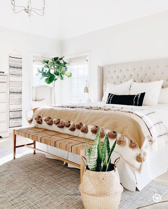 a chic neutral bedroom with a gorgeous creamy bed, a woven bench, some printed textiles and boho hangings on the wall
