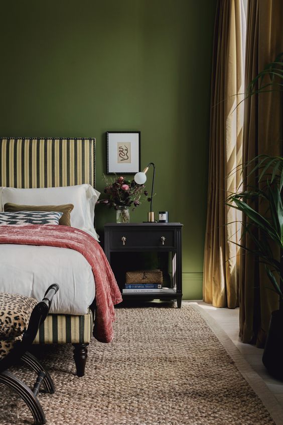 a chic vintage bedroom with green walls, a striped upholstered bed, printed bedding, a black nightstand and some decor