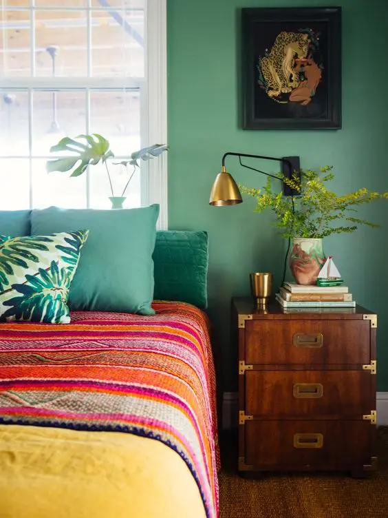 a colorful bedroom with green walls, a bed with bold bedding, a dark-stained nightstand, a gold sconce and some decor