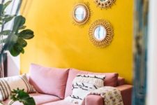 a colorful boho living room with a sunny yellow accent wall, a pink sofa, a gallery wall of mirrors and potted greenery