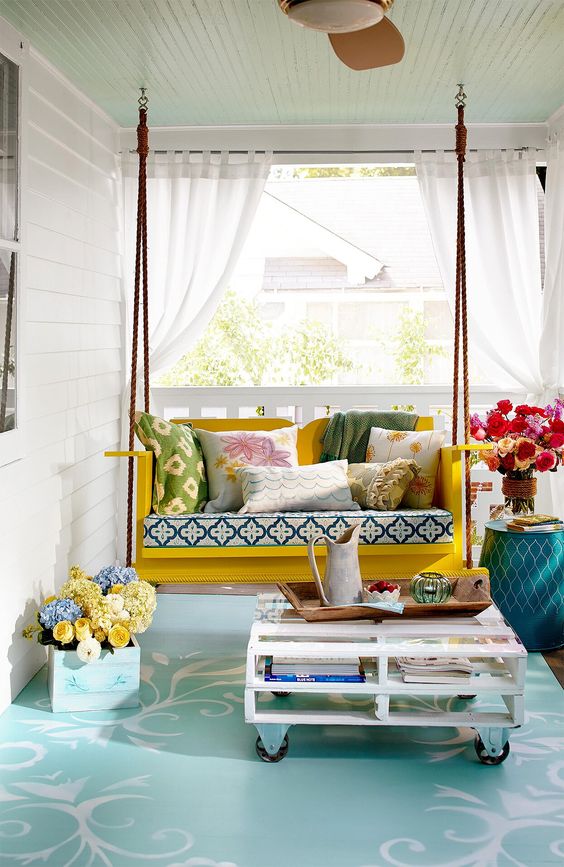 a colroful front porch with a yellow hanging bench, printed pillows, a pallet coffee table and some blooms