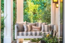 a cozy famrhouse porch with a hanging bench, striped upholstery, potted greenery and a wicker daybed is welcoming