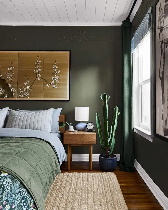a dark green bedroom with a bed with green and printed bedding, a stained nightstand, a woven rug and some artwork
