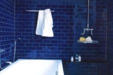 a deep blue bathroom fully clad with subway tiles, a white tub and neutral fixtures for a chic look