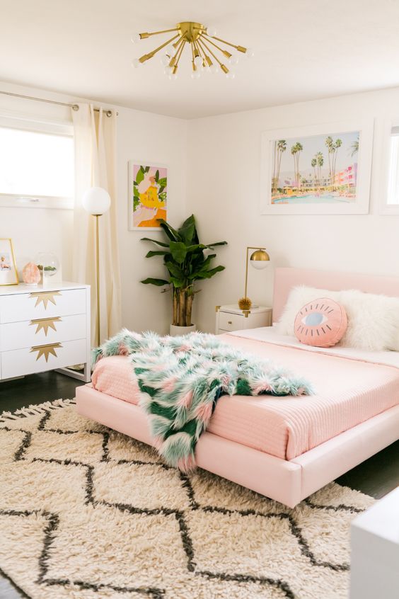 a glam tropical bedroom with a pink bed and bedding, an art deco dresser, a tropical artwork and a sunburst chandelier