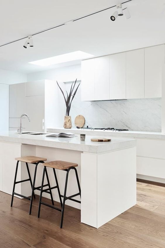a minimalist neutral kitchen with sleek cabinets, a white marble backsplash and countertops, woodne stools and a skylight