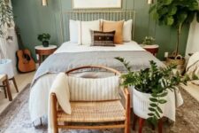 a modern bedroom with a sage green accent wall, a metal bed with neutral bedding, a woven chair, potted plants, gold touches for more elegance