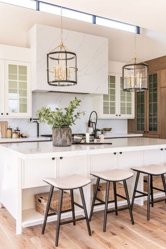 a modern neutral farmhouse kitchen with a white marble backsplash, countertops and a hood, glass uppers, pendant chandeliers