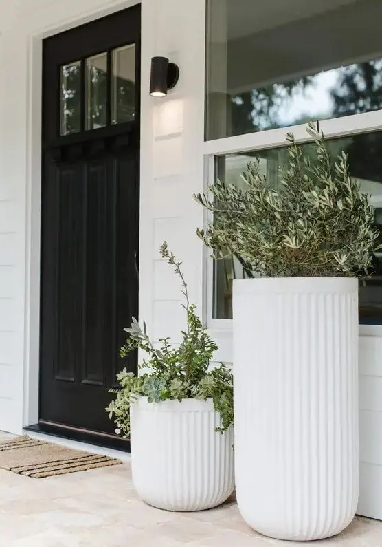 a modern porch with a black door, a duo of large fluted planters with greenery and a woven rug plus a black spotlight
